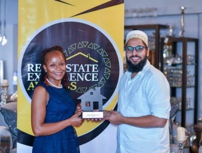 A woman and a man hold a house-shaped trophy in front of a Real Estate Excellence Awards banner